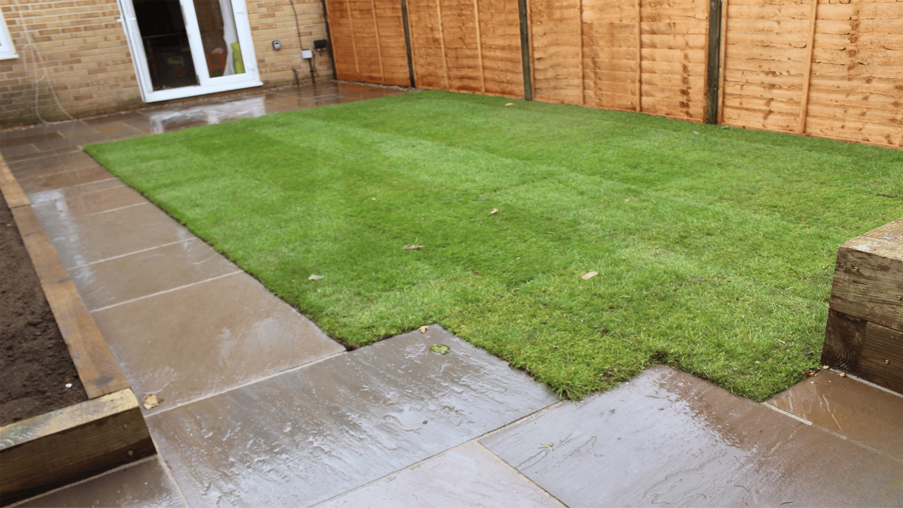 Day time backyard landscaping featuring modern Grey fence and white cushion chairs, low planter edge, stepping-stone pathway, and patio doors opening onto the garden.