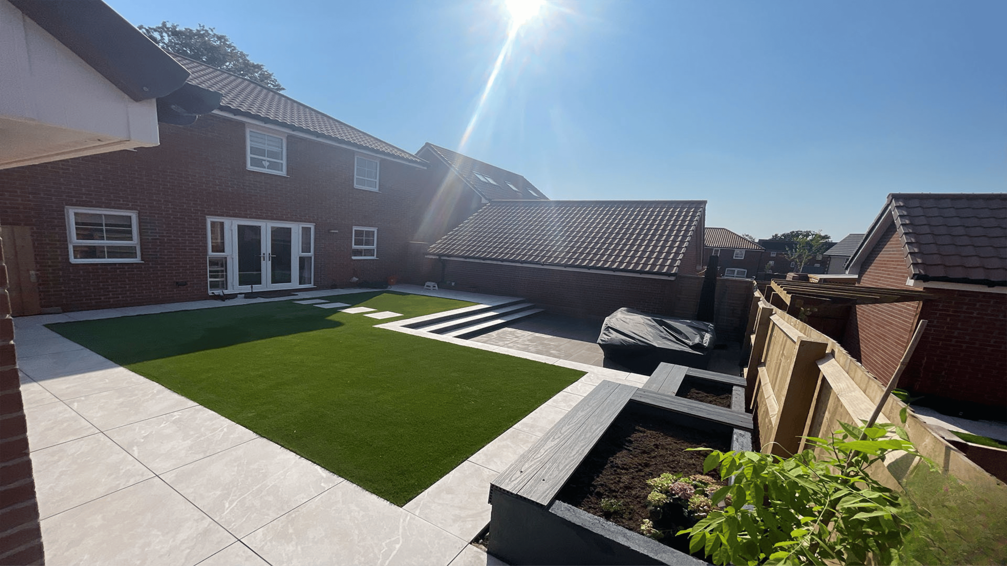 Modern backyard garden design featuring illuminated landscaping with turf lawn, white stepping stones, decorative pebbles, LED-lit trees and custom wooden seating area in Bridgwater